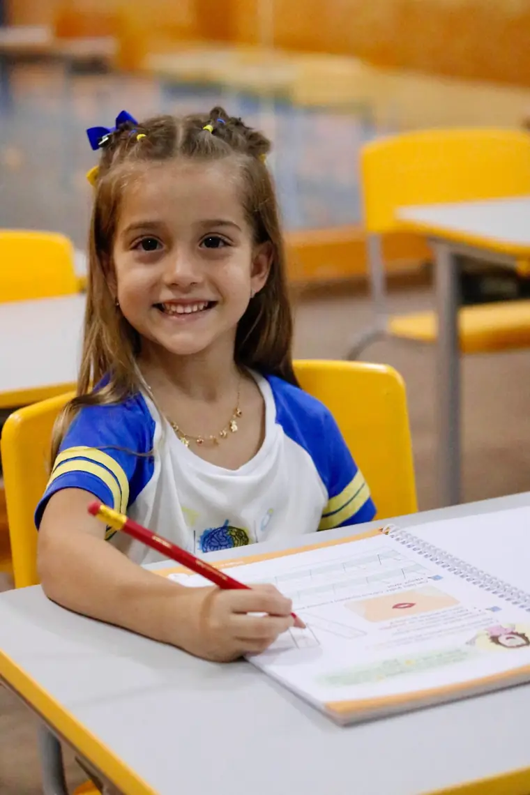 Menina de uniforme escrevendo a lápis numa apostila, à mesa da sala