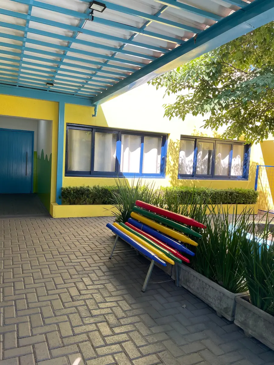 Entrada da escola: passarela coberta, prédio amarelo com esquadrias azuis e um banco de ripas coloridas