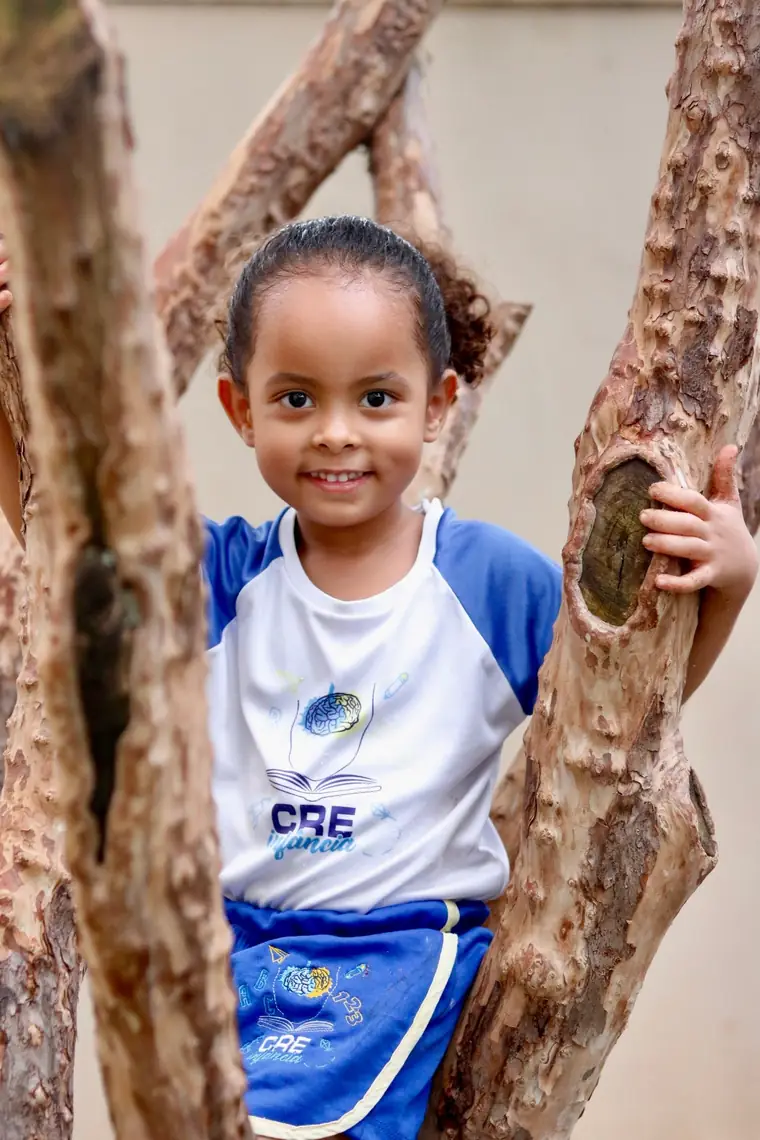 Menina de uniforme sorrindo junto a uma estrutura de troncos no quintal