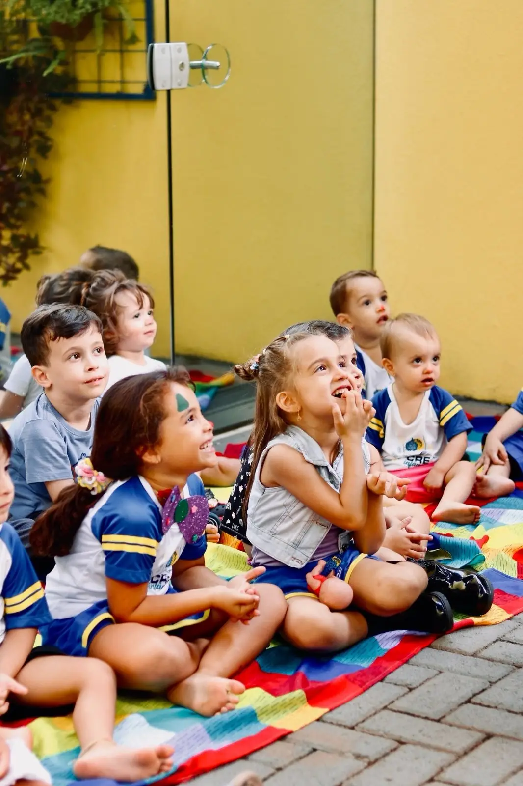 Turma de uniforme sentada num tapete colorido diante da parede amarela da escola